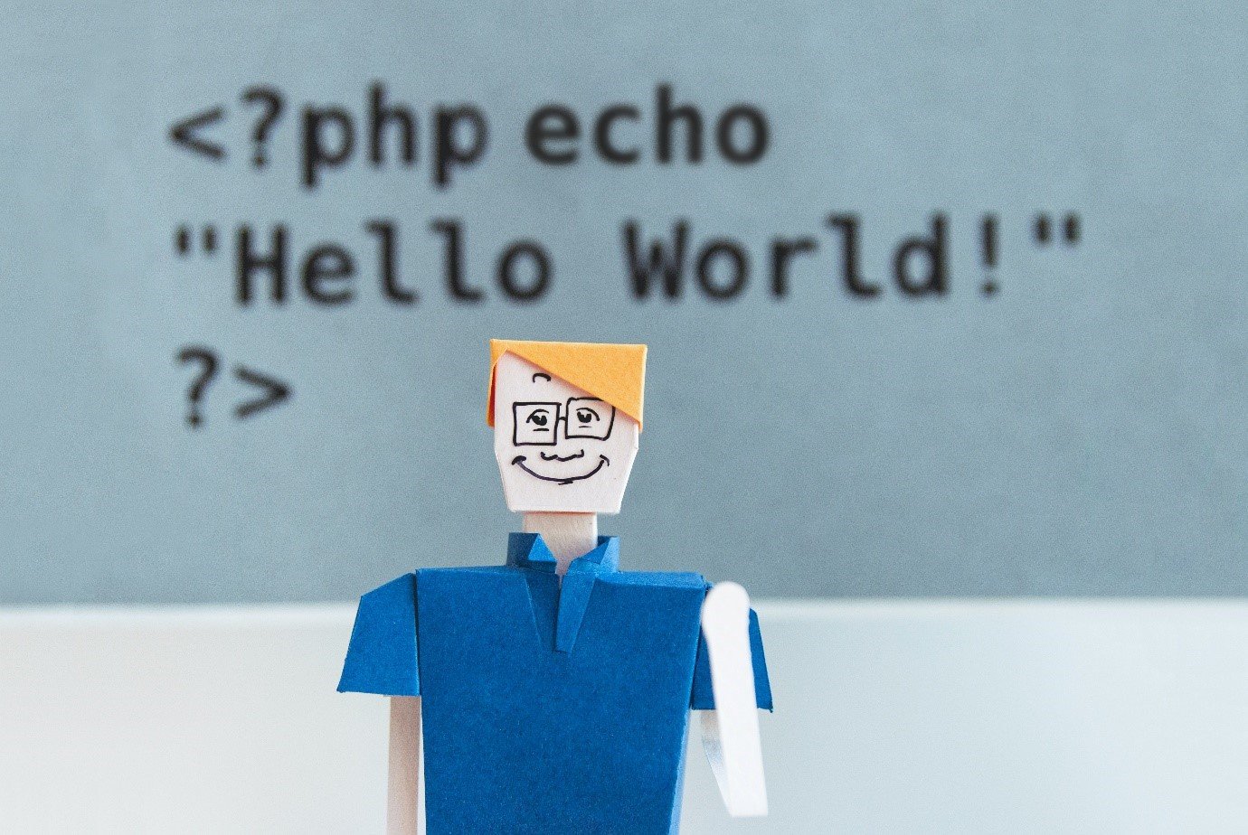 Paper figure in front of a screen showing a PHP "Hello World" echo statement